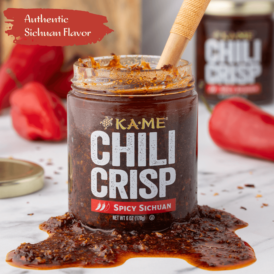 Jar of Kame Chili Crisp with a wooden spoon, surrounded by red peppers on a marble surface.