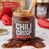 Jar of Kame Chili Crisp with a wooden spoon, surrounded by red peppers on a marble surface.
