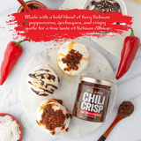 Kame Chili Crisp jar with decorative items on a white surface, featuring text about Sichuan peppercorns and garlic.