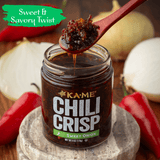 Jar of Ká-Mé Chili Crisp with a wooden spoon, surrounded by red peppers and onions, on a wooden surface.